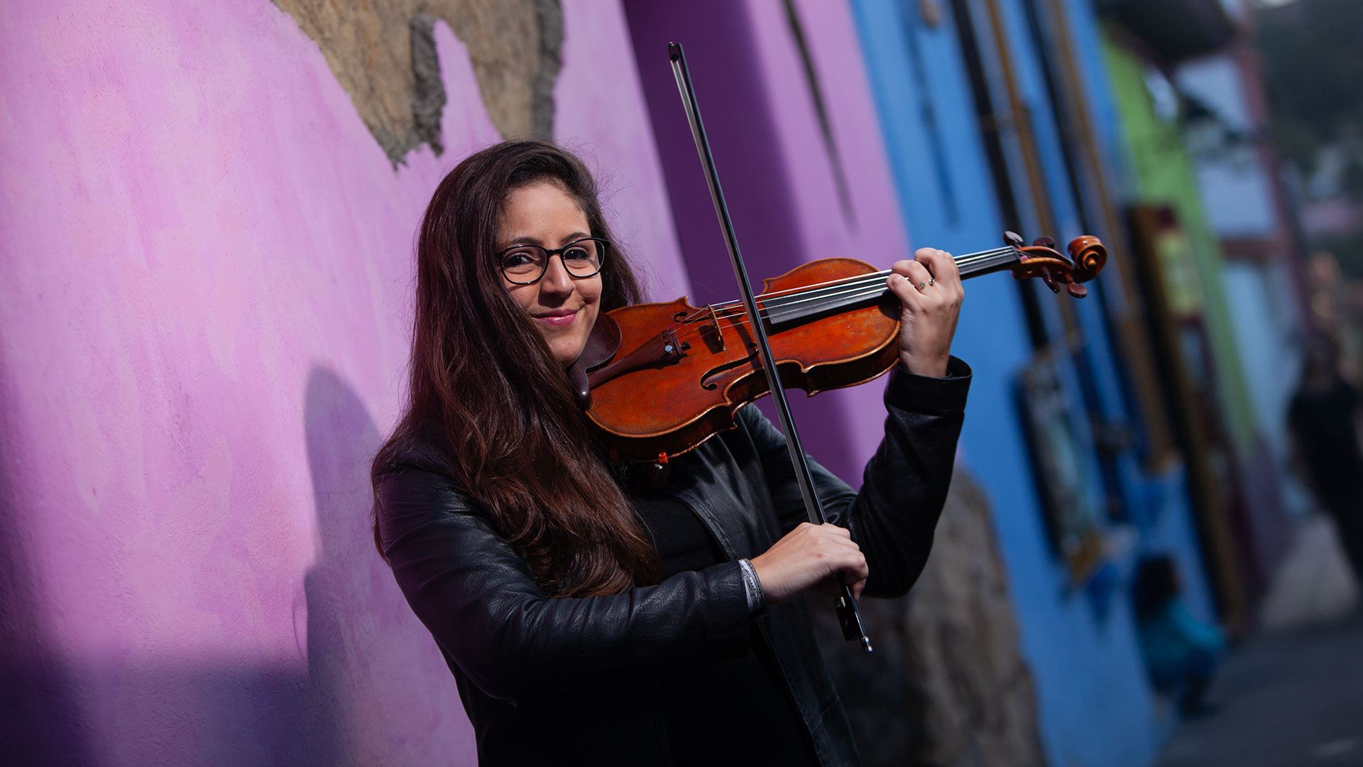 Daniela Padr&#243;n wearing a leather jacket and playing a violin in front of a colorful wall.